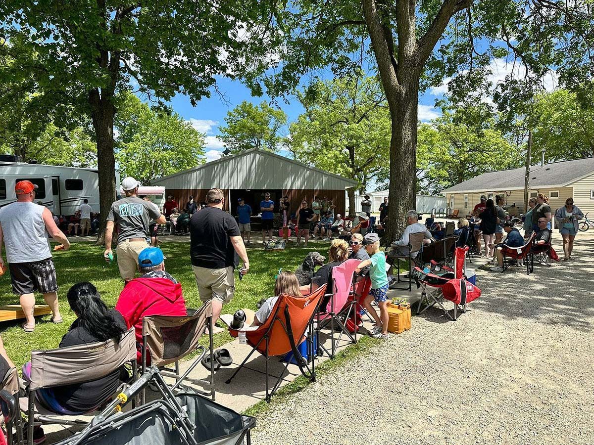 Outdoor gathering with people, chairs, and a small building under trees