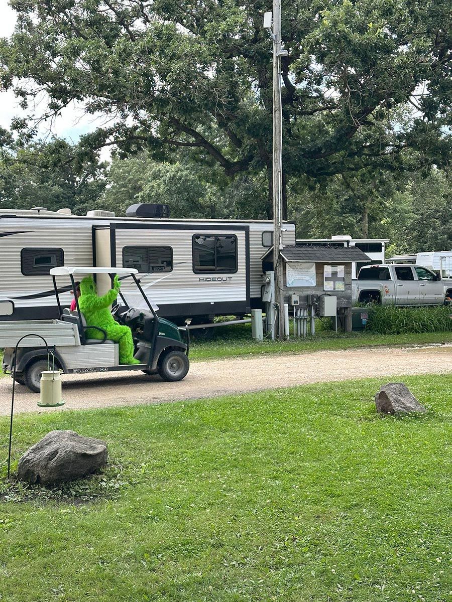 Person in green suit drives golf cart near a camper in a campground