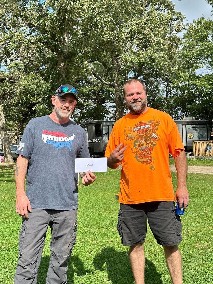 Two men outdoors: one in a cap and gray shirt, holding a paper, the other in an orange shirt