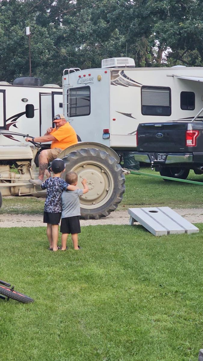 Man on tractor smiles at two children on a grassy lawn near an RV