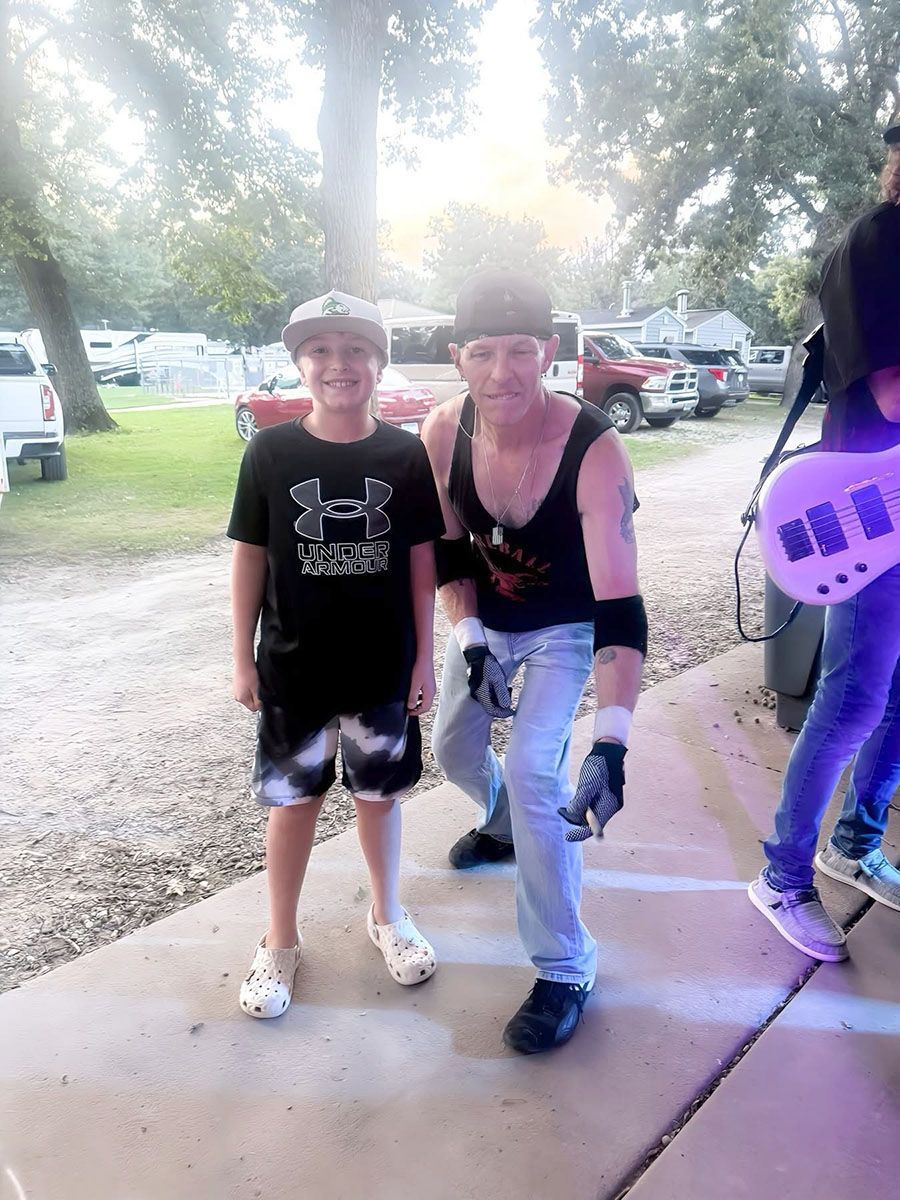 A boy in a cap and shorts stands with a man in a tank top, near a stage and cars