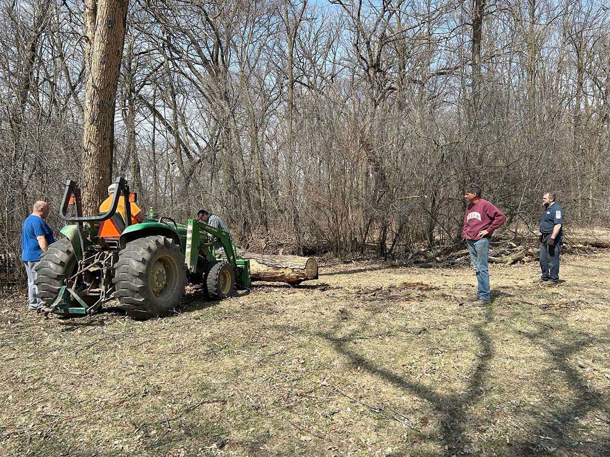 Three men watch a tractor with a log in a wooded area