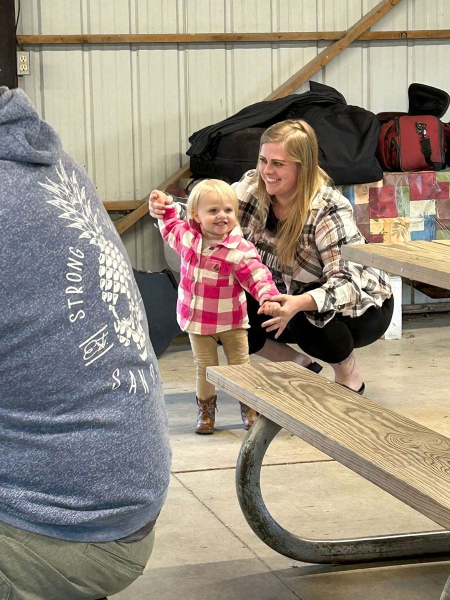 Woman sitting, holding hands with toddler wearing pink plaid shirt, near a picnic table