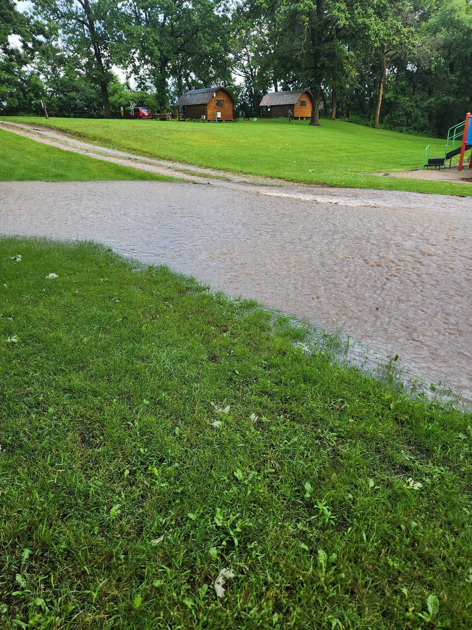 A dirt road going through a grassy field with a playground in the background.