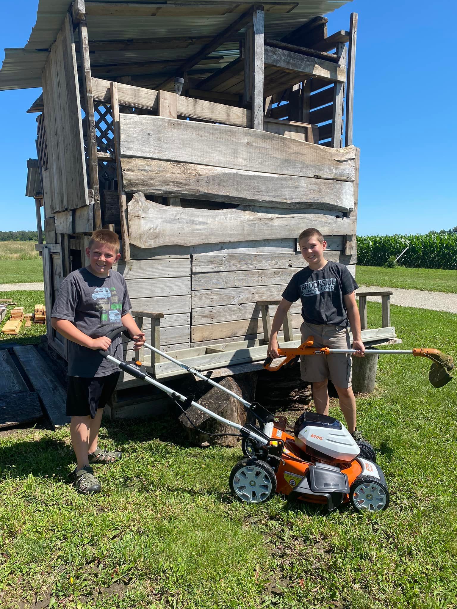 Two young men are standing next to a lawn mower in a field.