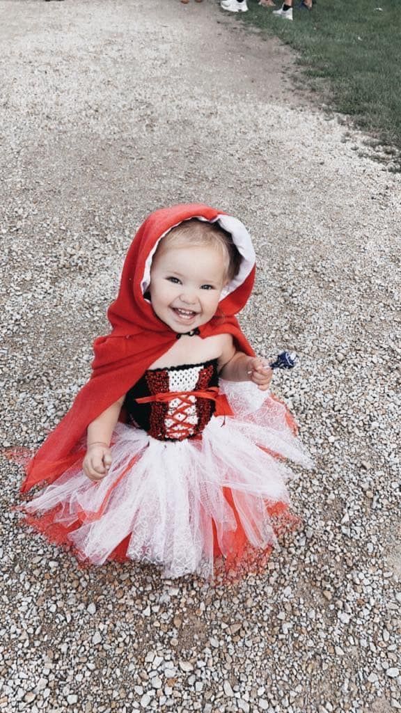 A little girl in a little red riding hood costume is sitting on the ground.