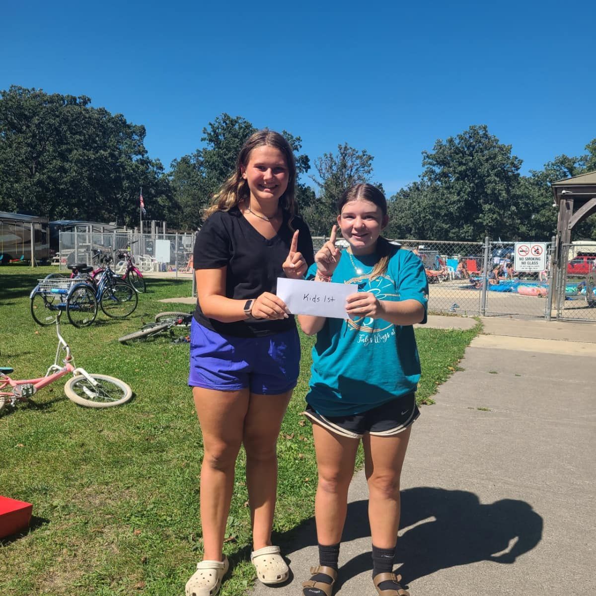 Two girls standing next to each other in a park holding a sign.