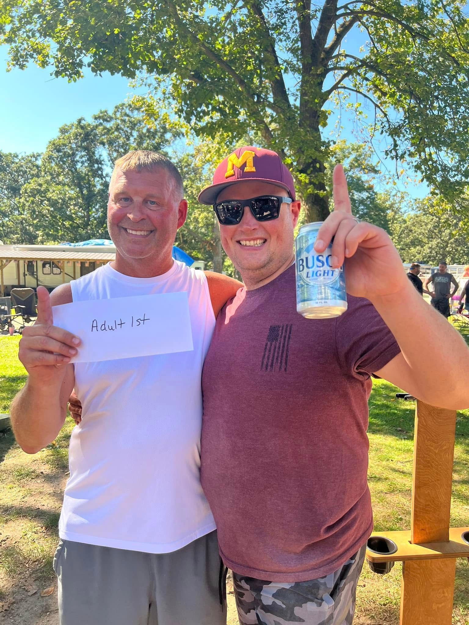 Two men are standing next to each other in a park holding a can of beer.