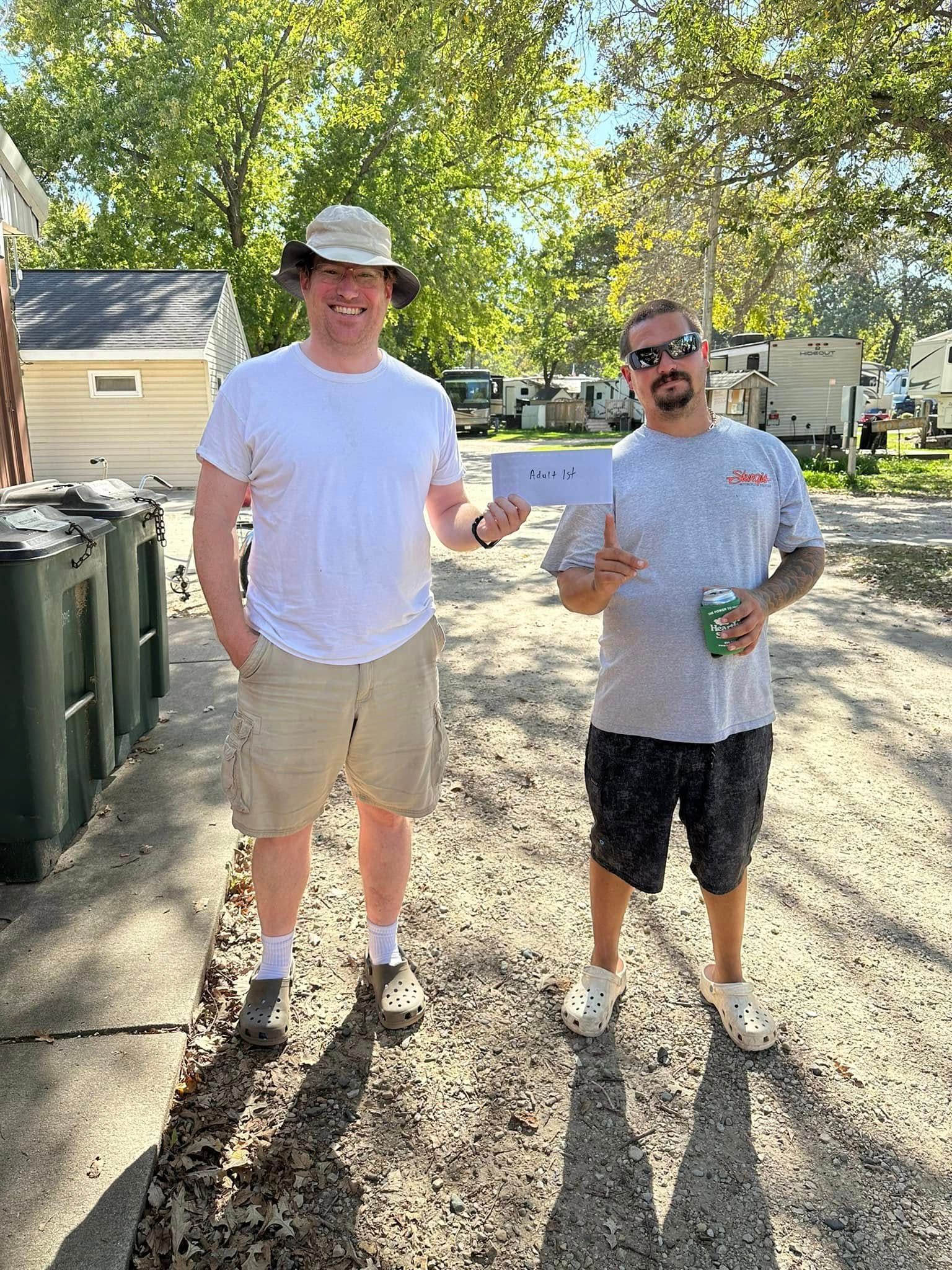 Two men are standing next to each other on a dirt road holding a check.