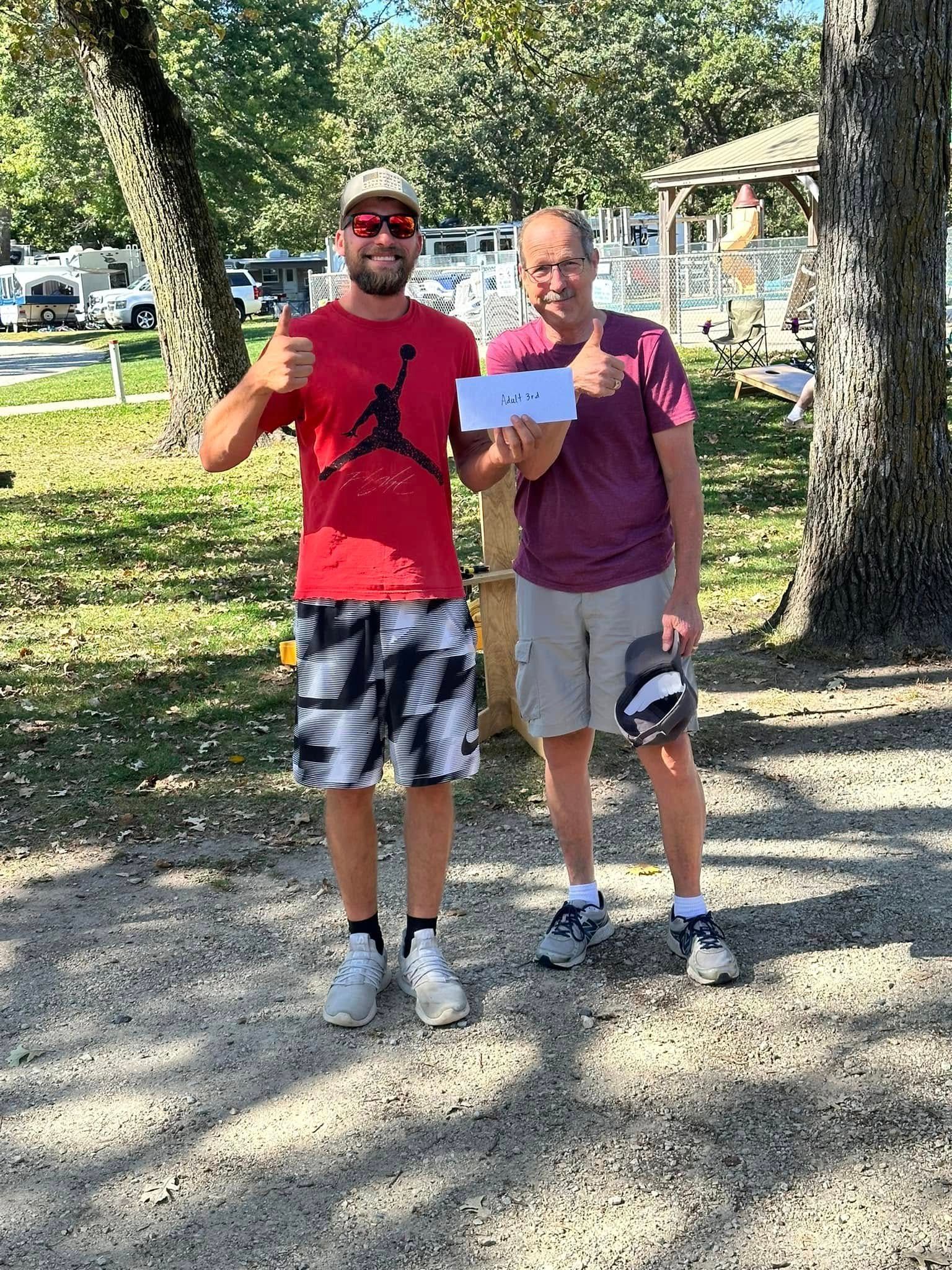 Two men are standing next to each other in a park holding a paper and giving a thumbs up.
