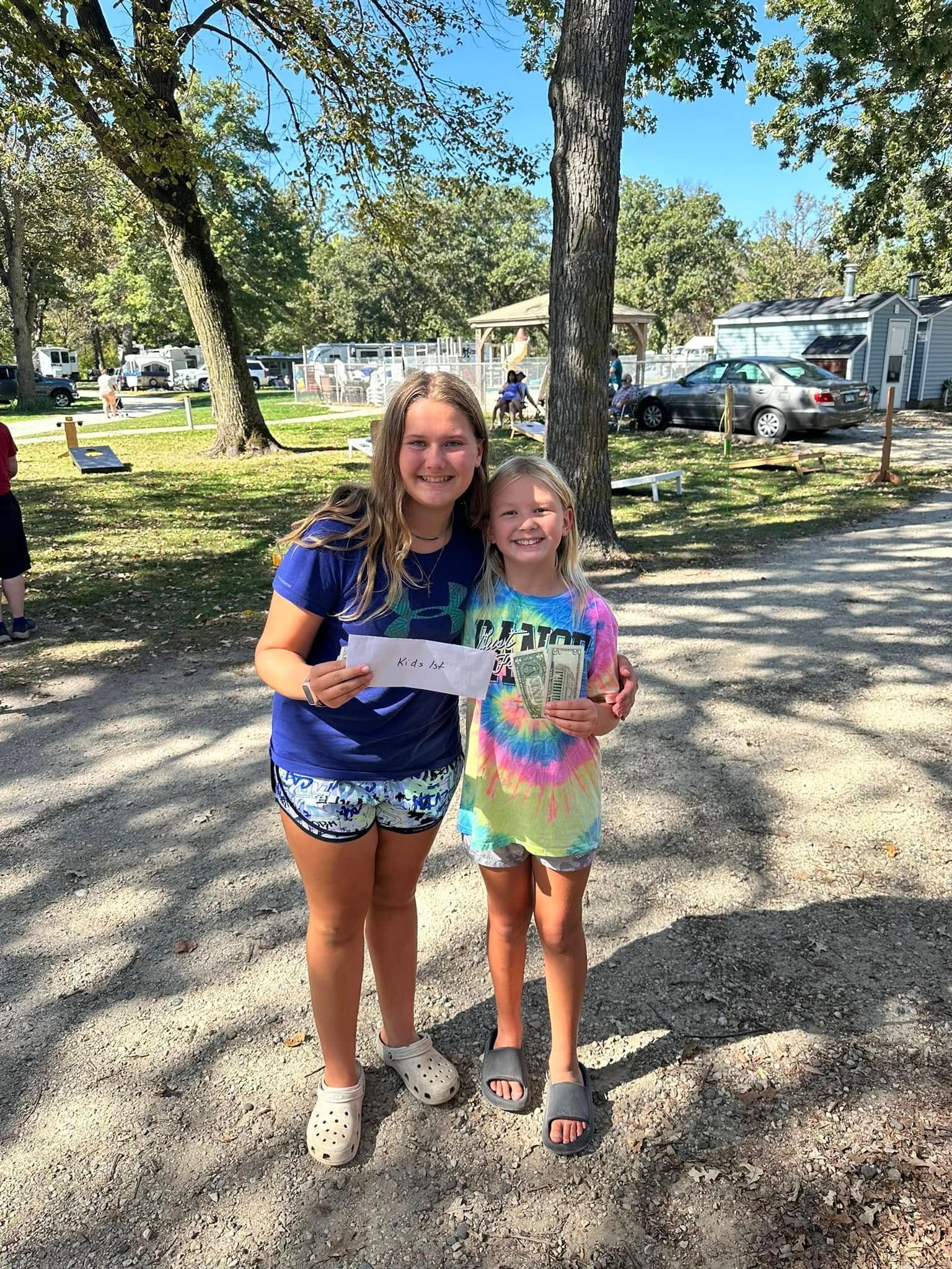 Two young girls are standing next to each other in a park holding a paper.