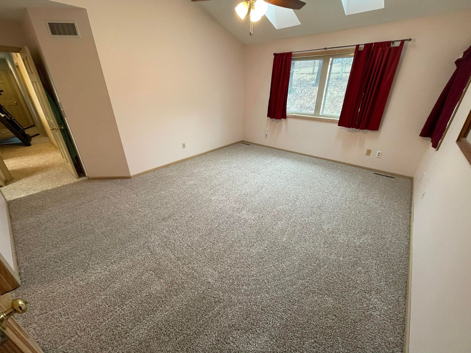 Empty room with pink walls, textured carpet, window with red curtains, and open door.