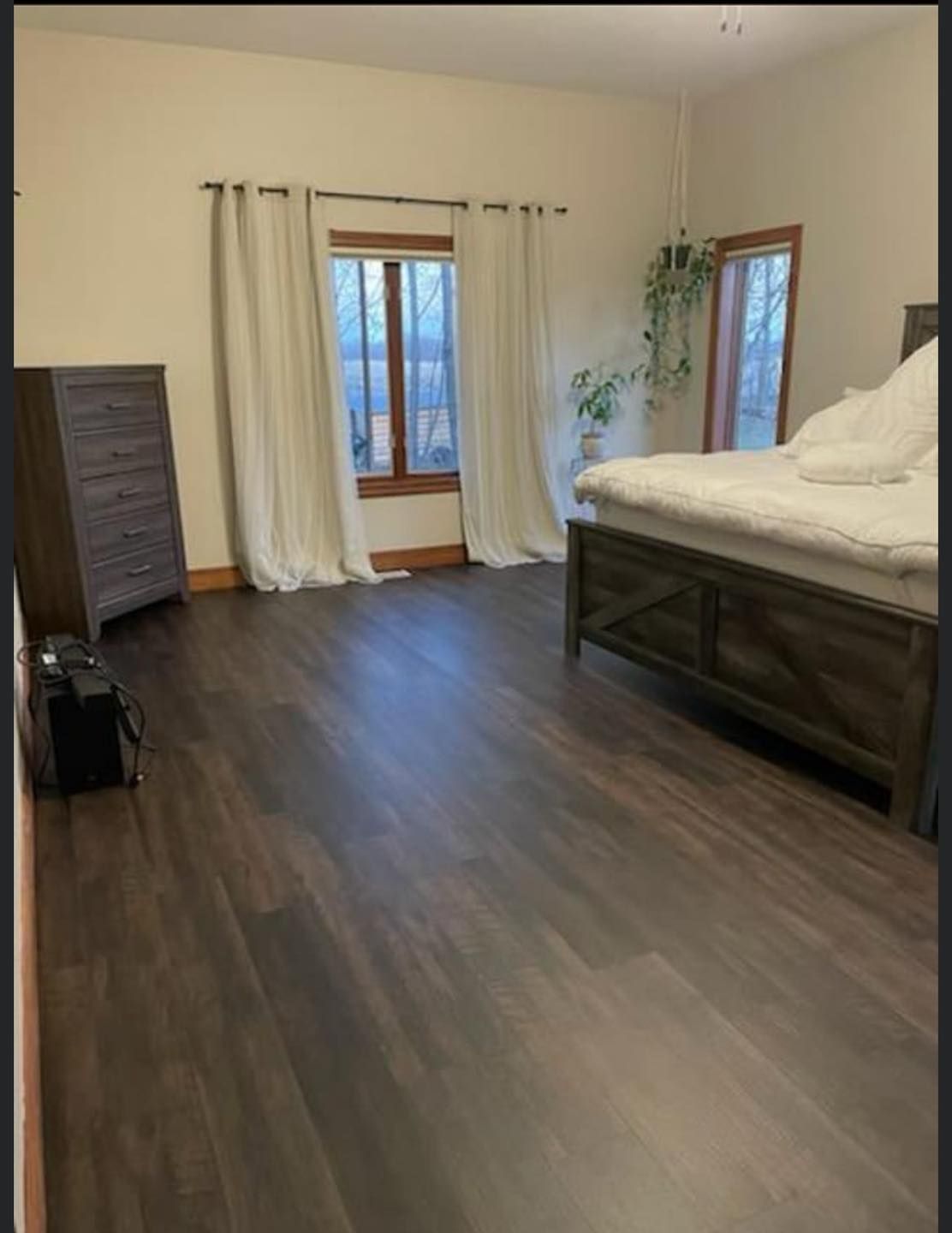 Bedroom with dark wood flooring, wooden furniture, white curtains, and windows.