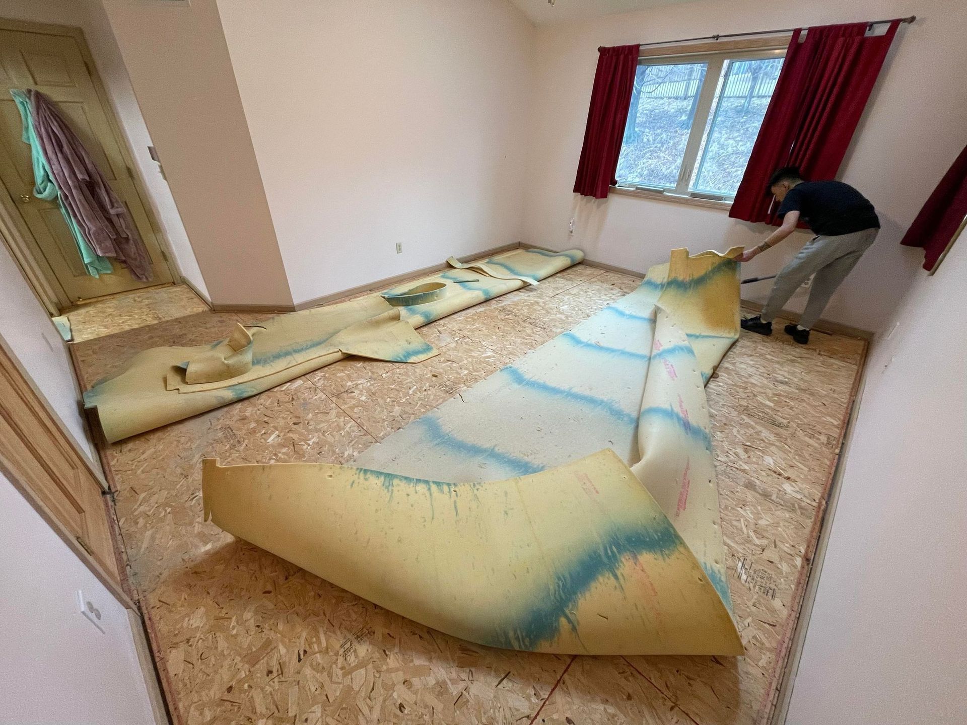 Man removing carpet in a room with bare wooden flooring and a window.