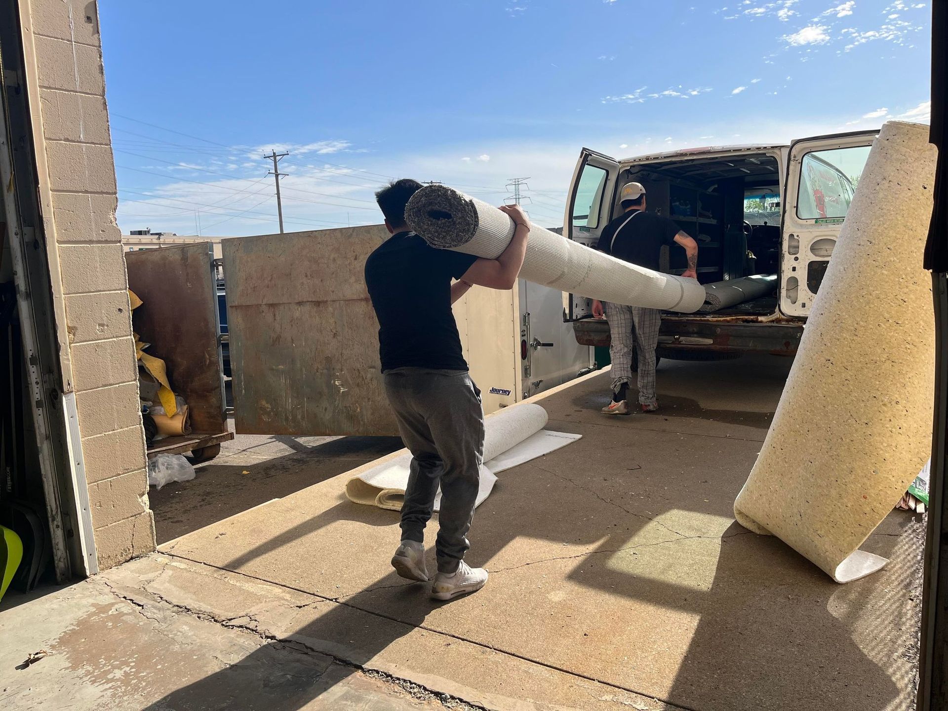 Two people loading rolled carpet into a van, under a clear sky.