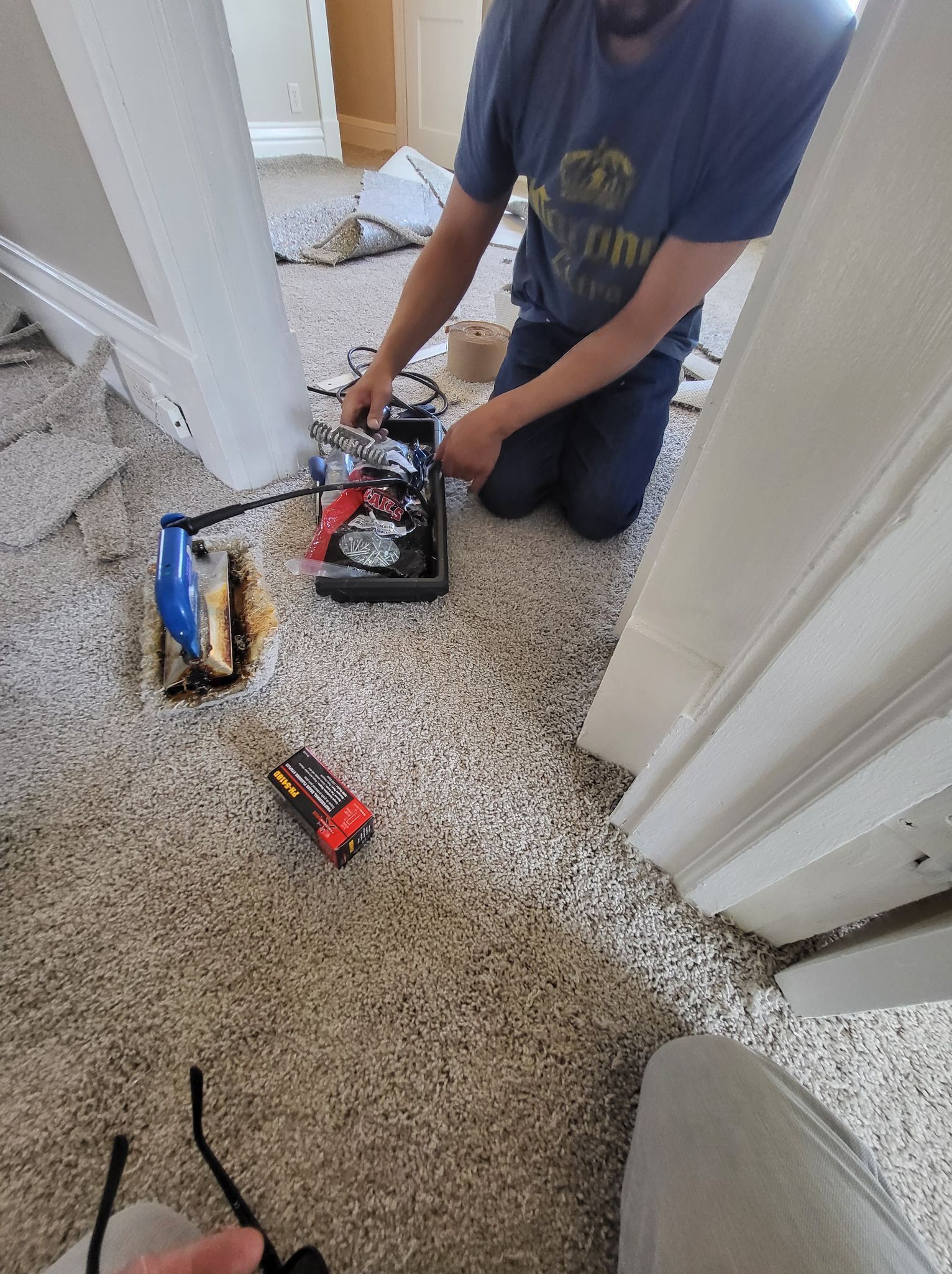 Person kneeling, holding tools, near a carpeted doorway. A toolbox, a small car, and a light are visible.