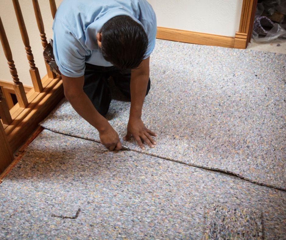 Person kneeling, cutting carpet on a floor near wooden stairs; a construction scene.