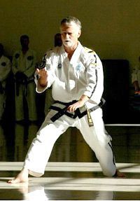 A man in a white karate uniform with a black belt is kneeling on the floor.