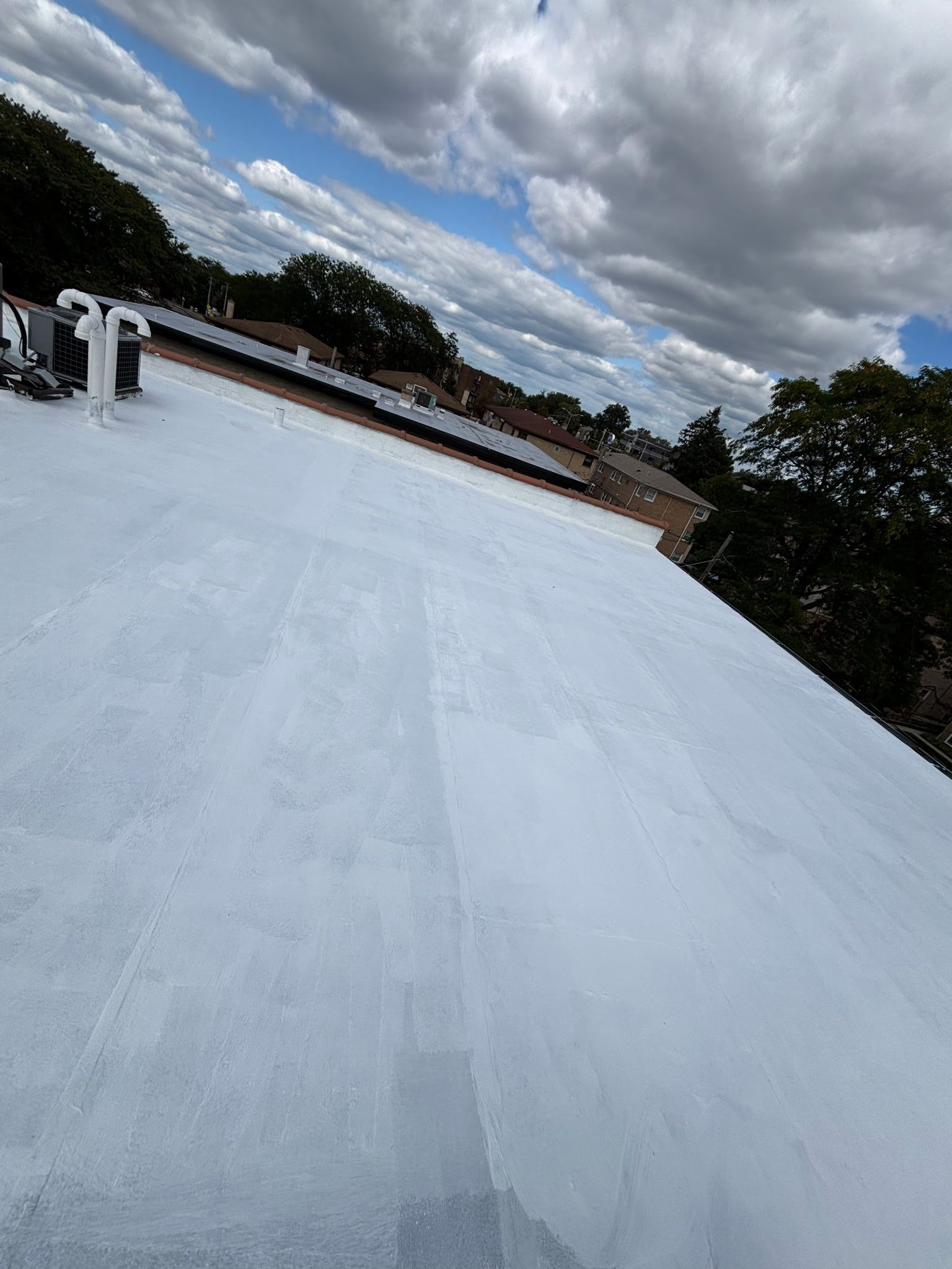 White-coated flat roof under a cloudy sky. Trees and rooftops visible in the distance.