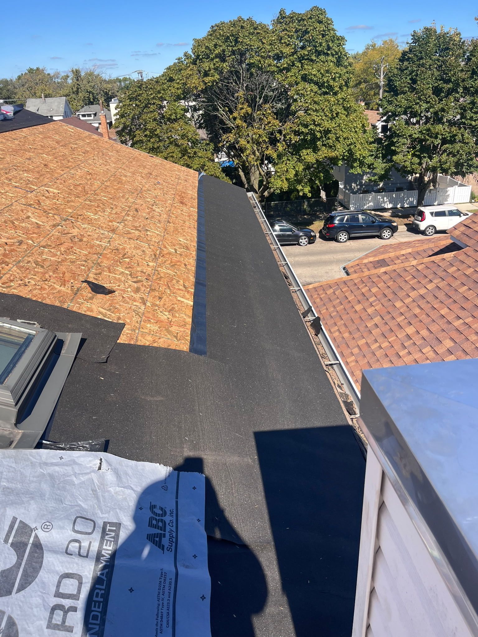 View from roof edge with new black roofing meeting aged brown shingles; street with cars in background.
