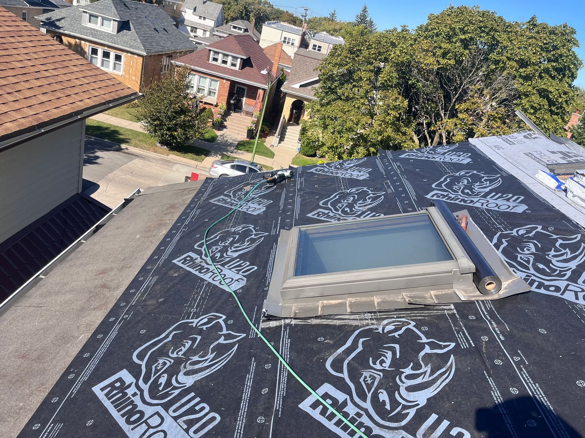 Rooftop view with a skylight and RhinoRoof underlayment. Houses and trees in the background under a blue sky.