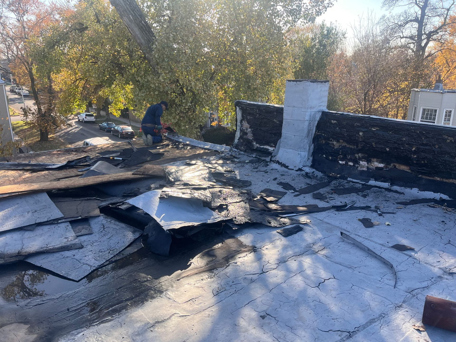 Roofer working on a flat roof with torn, dark roofing material. Sunlight streams through trees in the background.