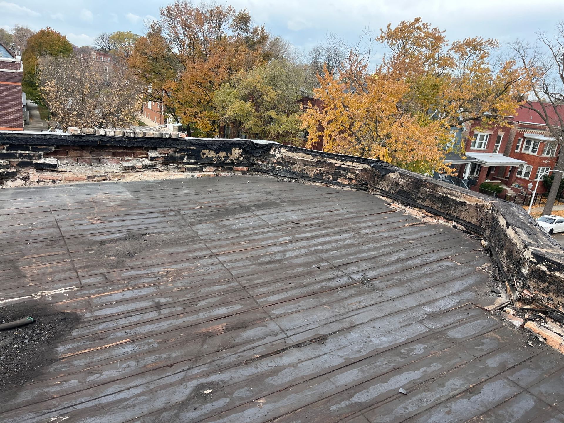 Damaged rooftop with exposed boards and charred edges; trees and houses in the background.