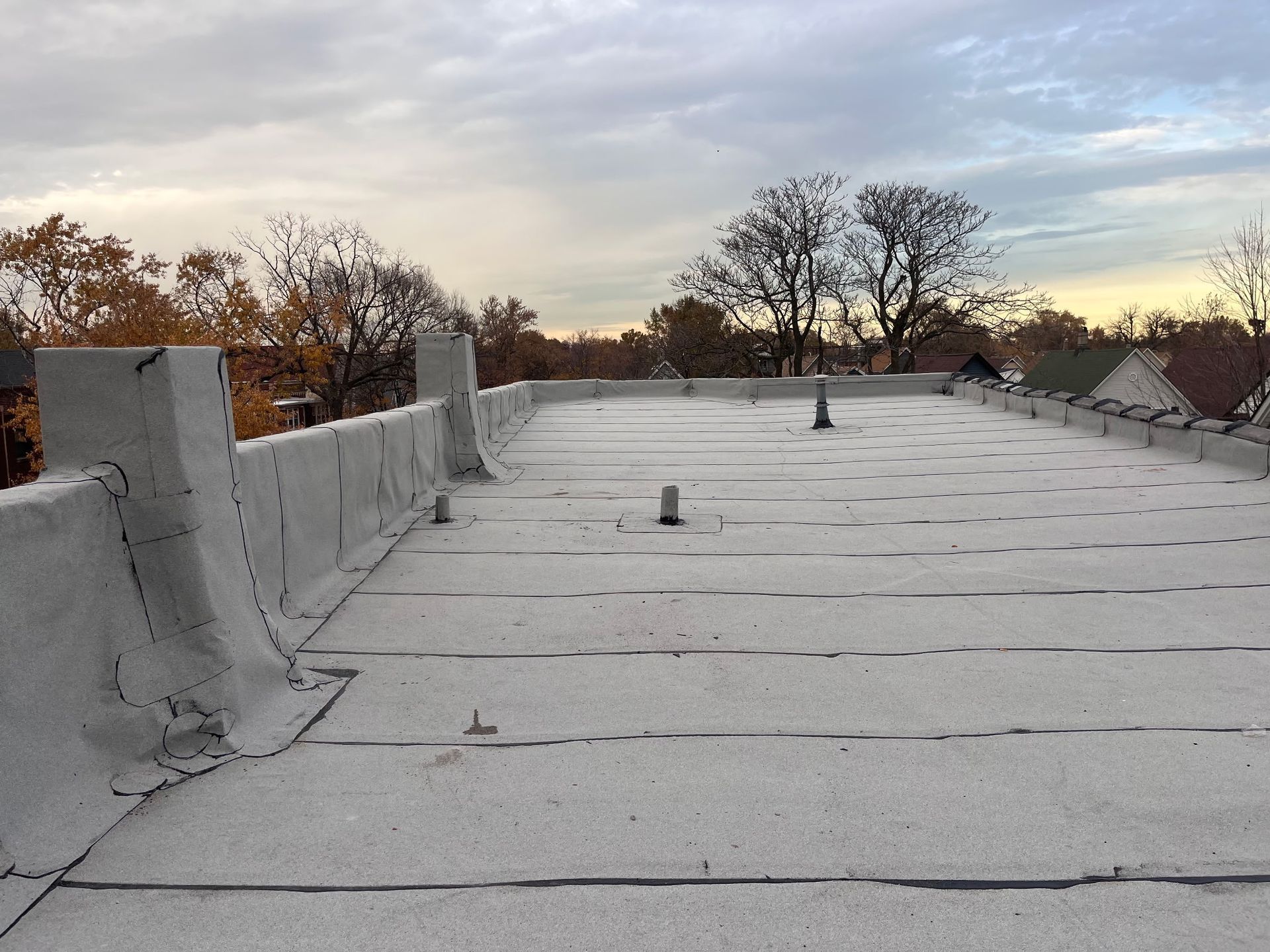 Flat rooftop with a gray surface and perimeter walls, cloudy sky in the background, trees in the distance.