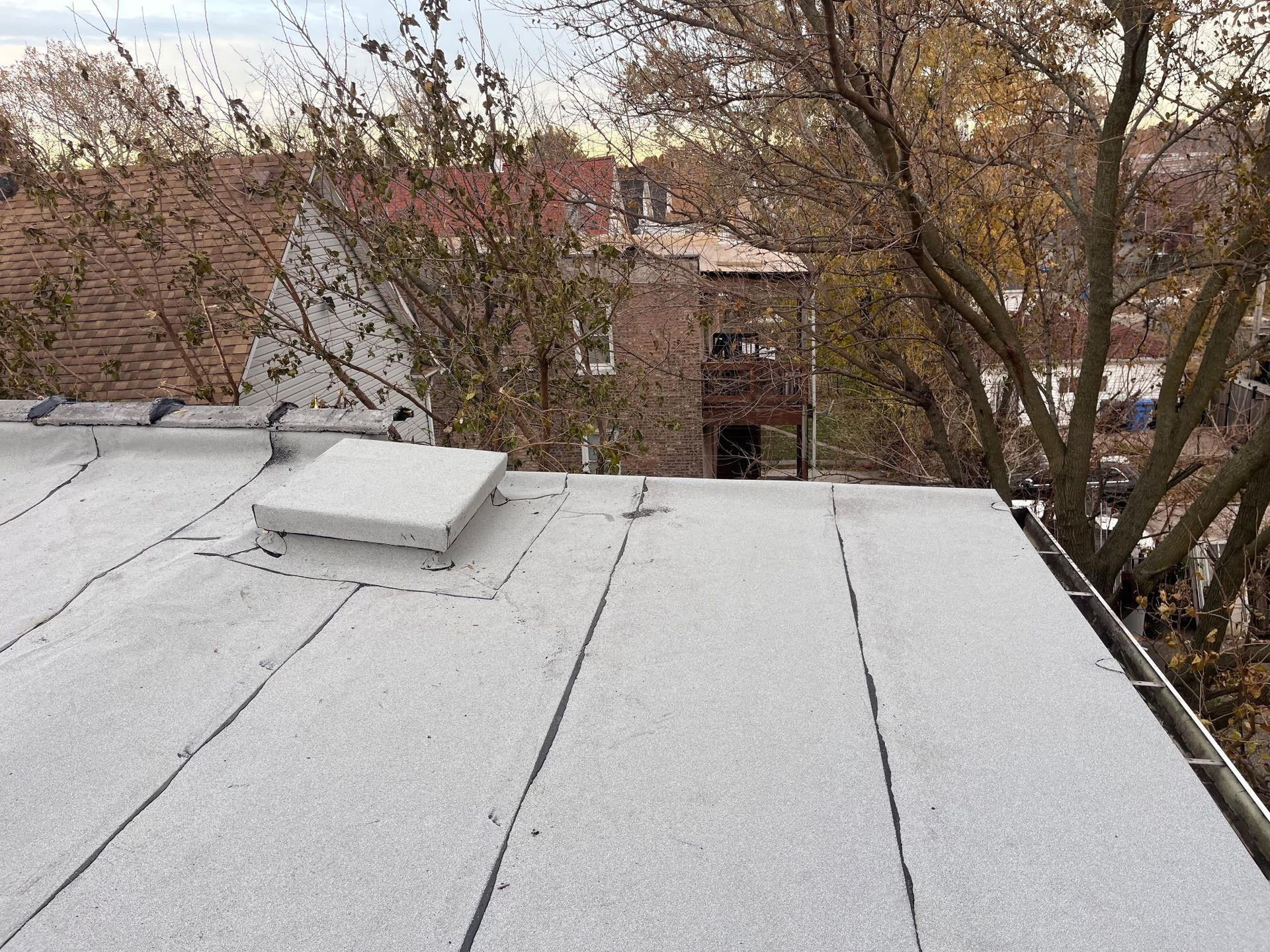 Flat rooftop with white gravel surface and a rectangular vent, with a brick building and trees in the background.