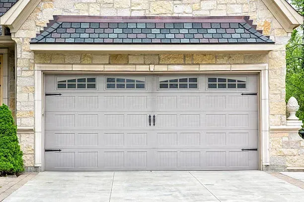 A closed, light-gray carriage-style garage door with four windows and decorative black hinges on a stone-faced house.