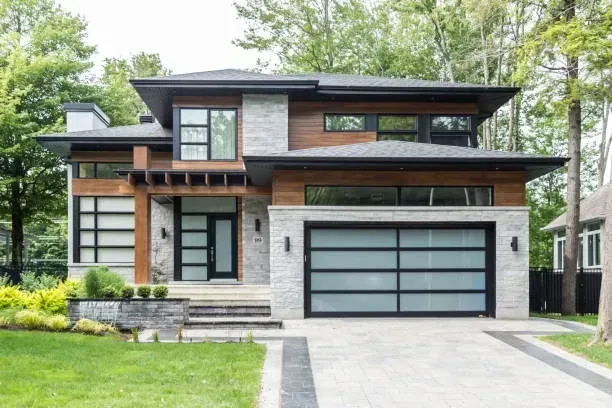 A modern two-story home with light stone, wood siding, large windows, and a frosted glass garage door in a wooded setting.