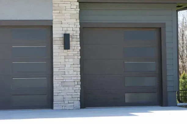 Two modern, dark gray garage doors with rectangular glass windows, separated by a light stone pillar.