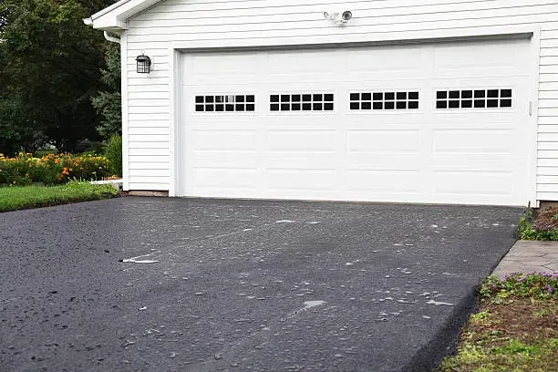 A white garage with a closed door and four rows of windows above a newly paved asphalt driveway.