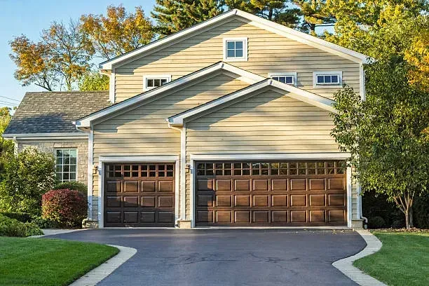 A beige, two-story house with a three-car garage, dark brown doors, and a paved driveway surrounded by trees and lawn.