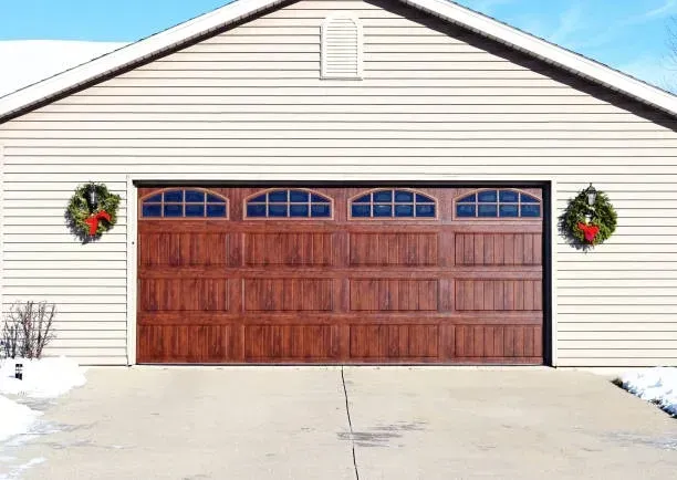 A beige garage with a wood-grain door and festive wreaths on each side, situated on a snowy concrete driveway.