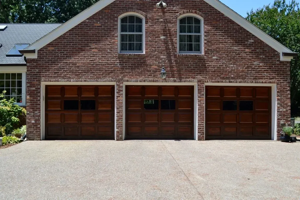Three-car garage with wood doors and a red brick exterior under a gabled roof, viewed from a gravel driveway.