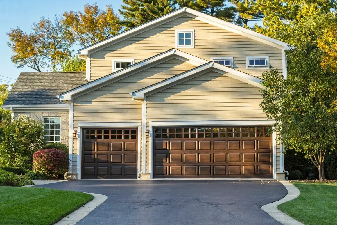A two-story tan suburban house with two dark brown garage doors, a long driveway, and surrounding trees.