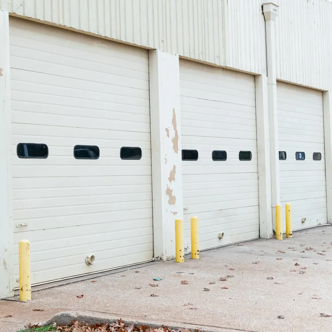 Three cream-colored, sectioned industrial roll-up doors with horizontal windows, set in a building with yellow bollards.