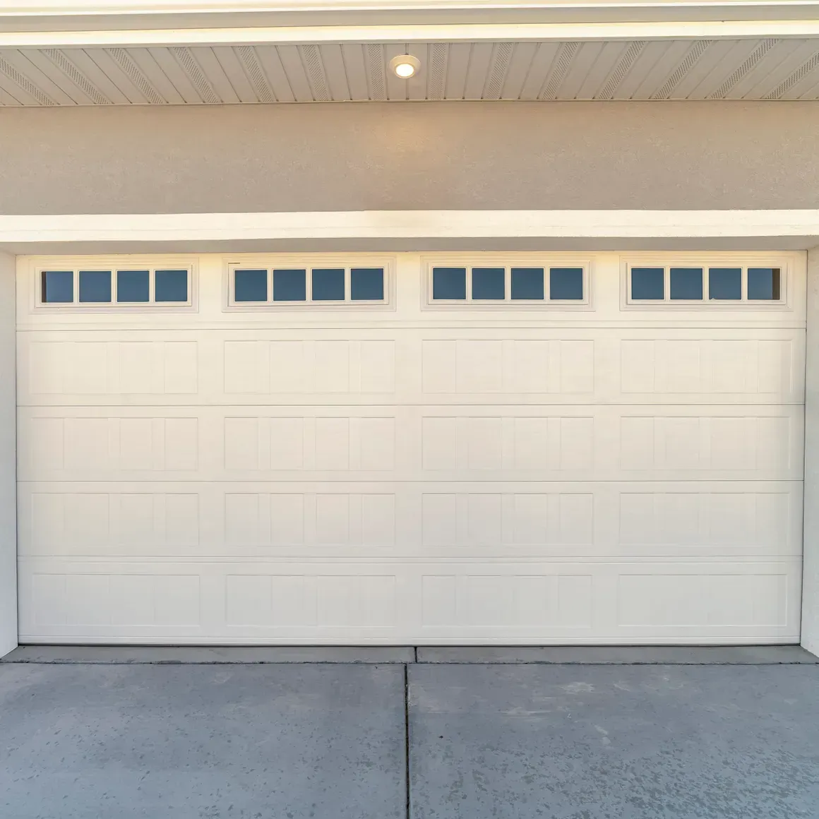 A white, four-paneled garage door with a row of four rectangular windows at the top, installed in a concrete driveway.