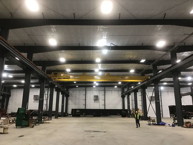 Large empty warehouse with steel beams, bright overhead lights, and one worker near the center.