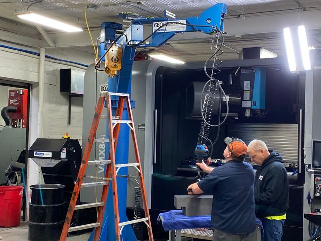 Two workers inspect a blue overhead hoist in a workshop with ladders and tools.