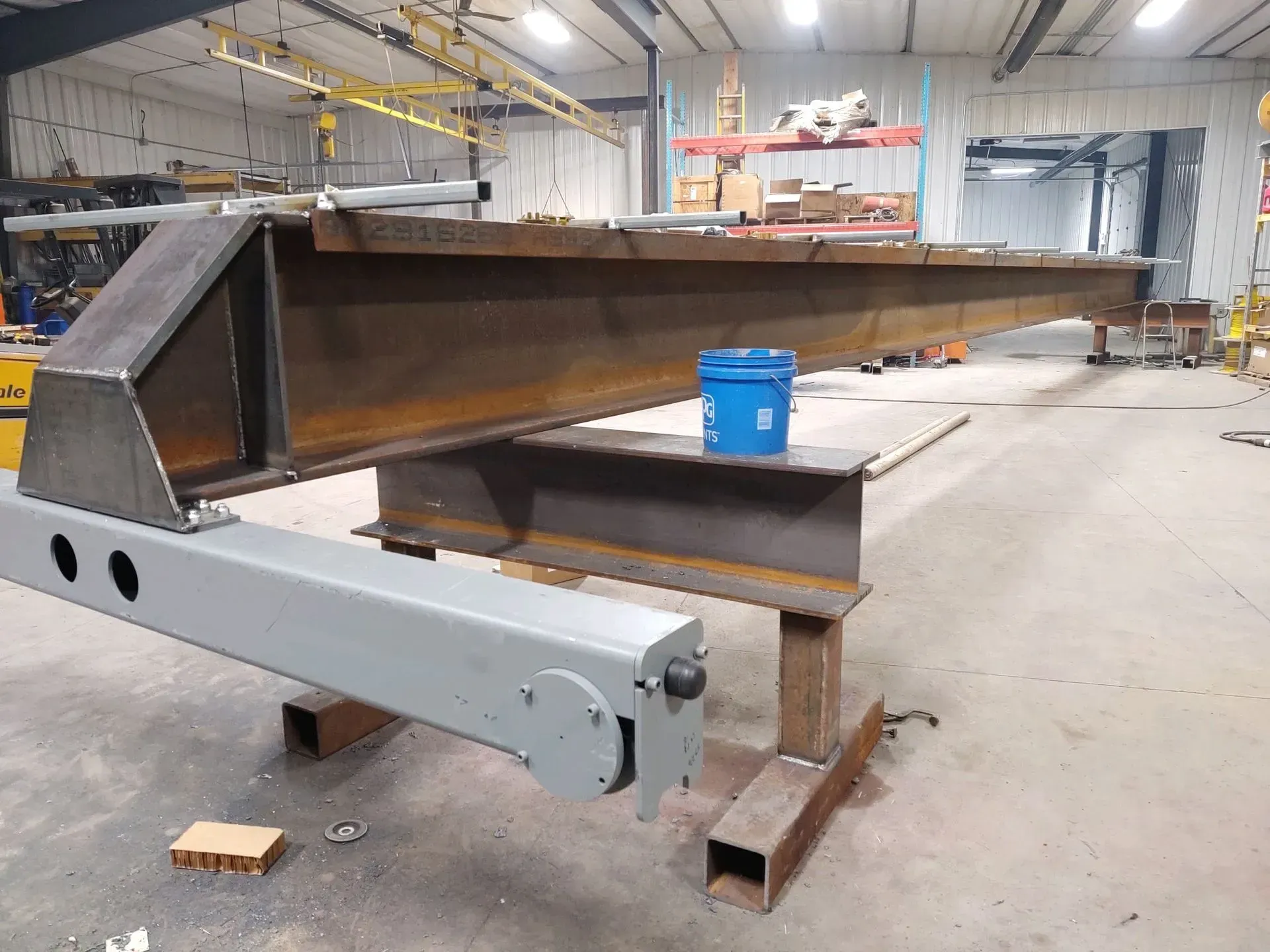 Large steel beam structure in a workshop, with a gray support arm and blue bucket nearby.