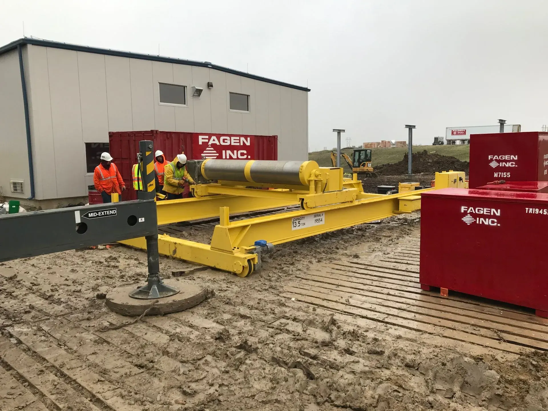 Workers in hi-vis gear beside yellow and red industrial equipment at a muddy construction site.