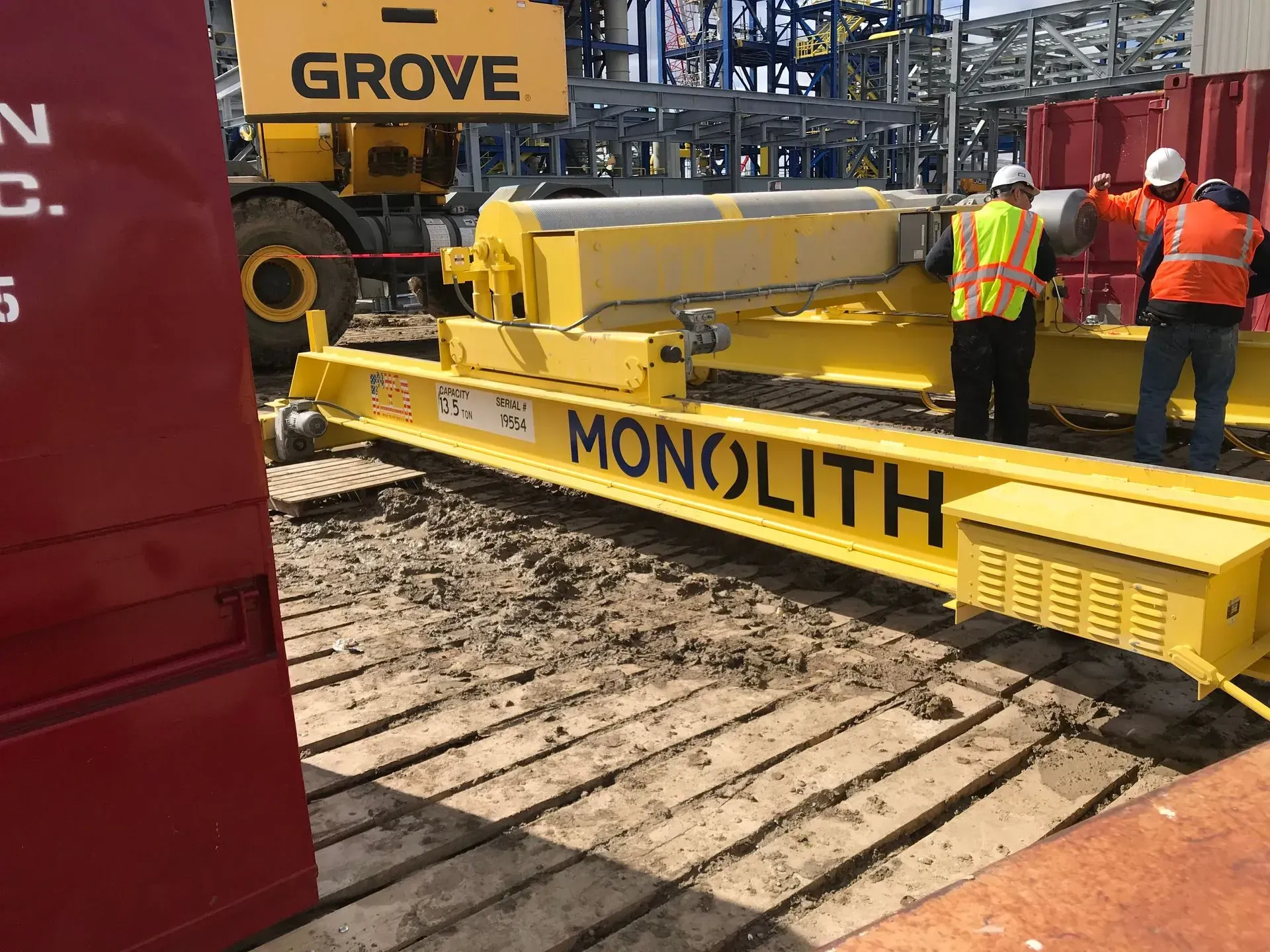 Yellow Monolith construction machine on muddy site with workers in orange safety gear and a Grove crane nearby.