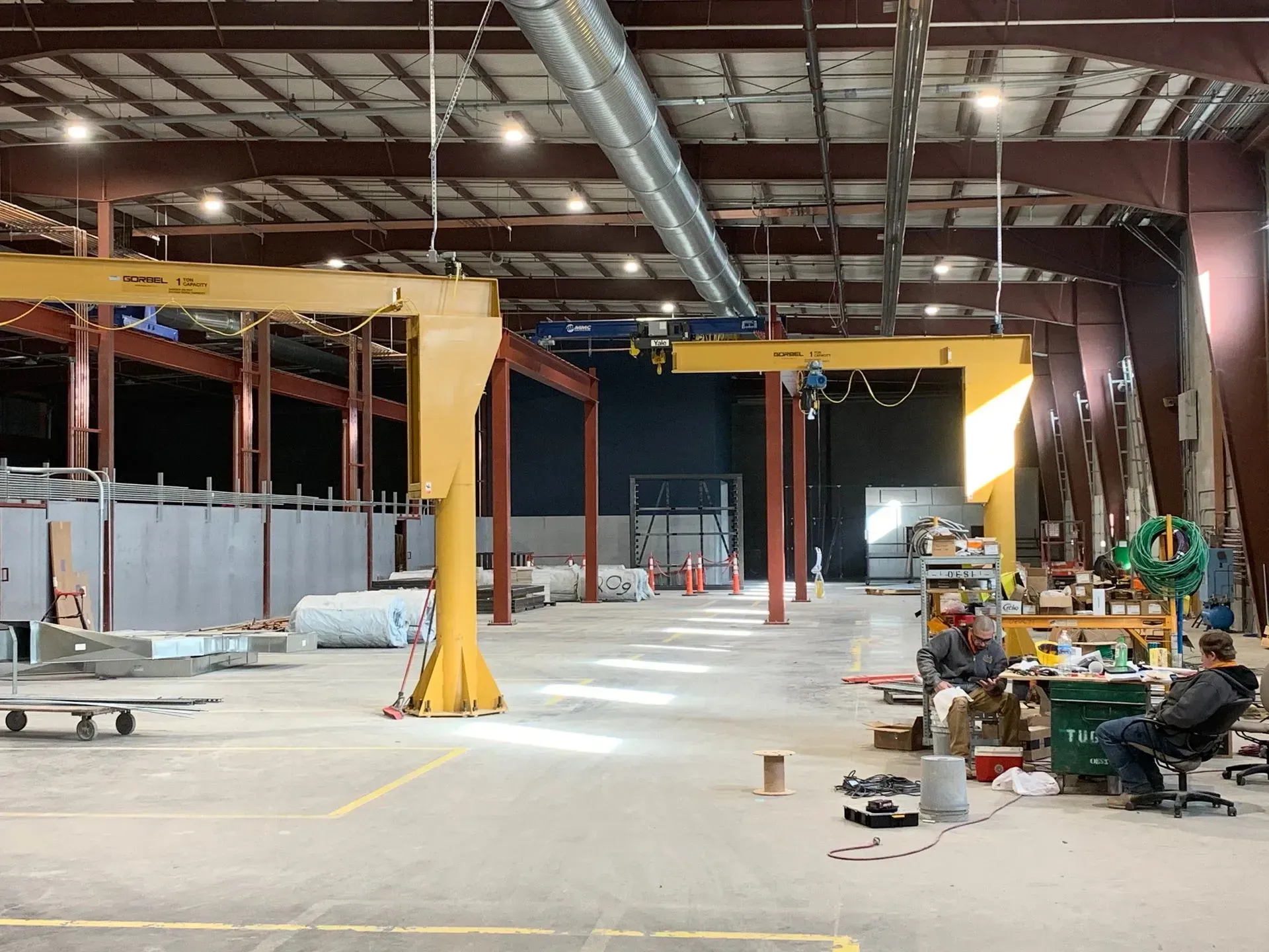 Large industrial warehouse interior with overhead cranes, equipment, and construction materials scattered on the floor.