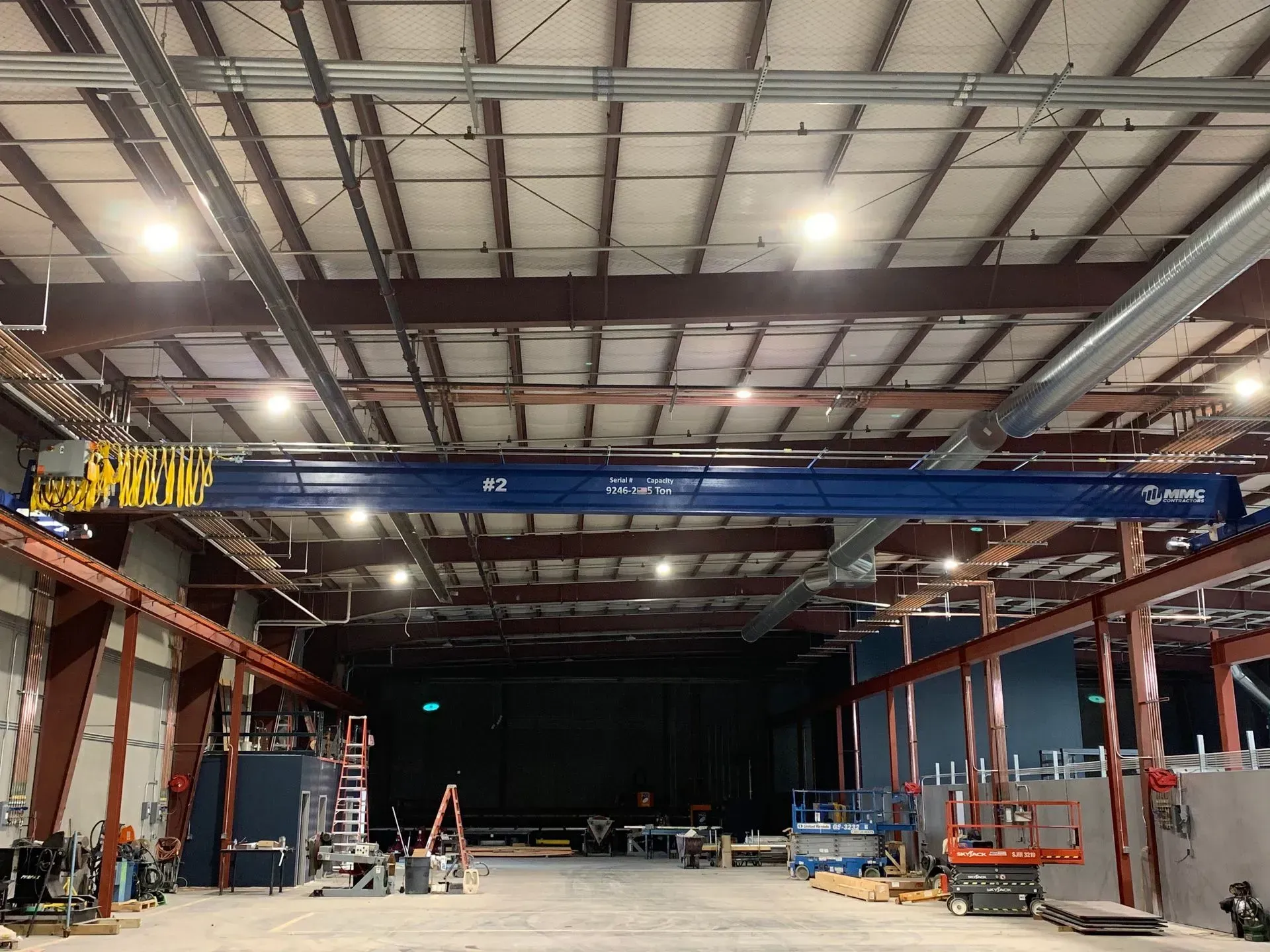 Large warehouse interior with steel beams, overhead crane, and ladders under construction lights.