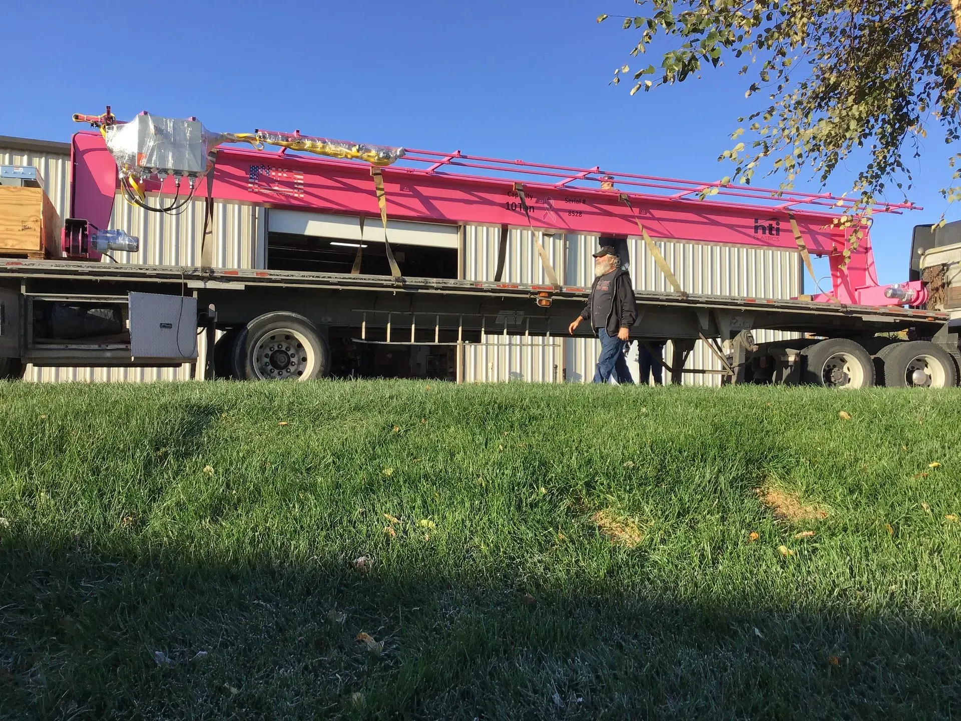 Flatbed truck hauling a long red crane beam past a grassy roadside, with one person walking beside it under a blue sky.