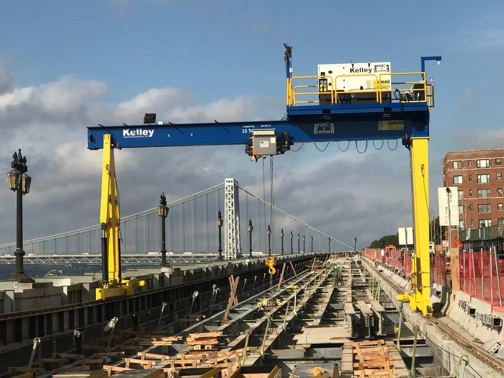 Rail-mounted crane over a bridge under construction with cables and city buildings in the background.