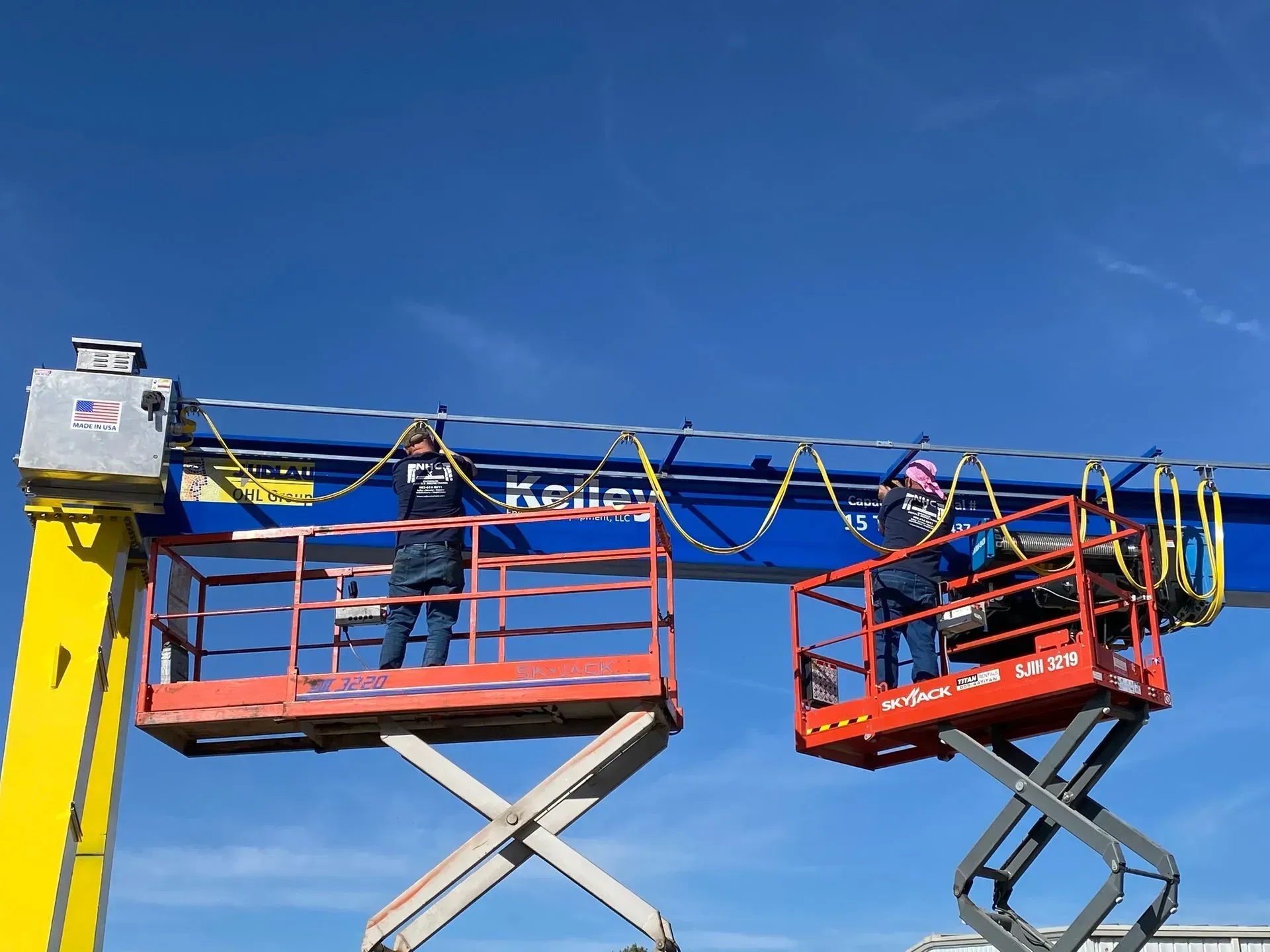 Two scissor lifts with workers under a blue sky, connected by a banner-like cable setup.