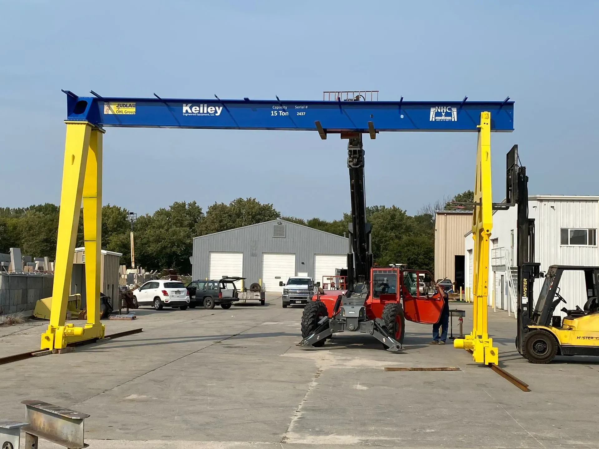 Yellow and blue overhead crane in an industrial yard with forklifts and equipment nearby.